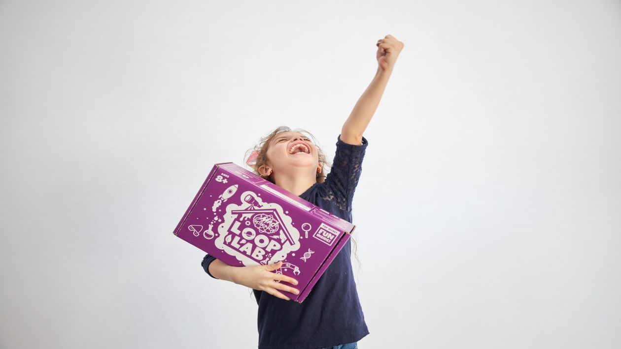 A very excited girl with one hand holding a Mad Science Loop Lab subscription box while the other hand is raised in the air
