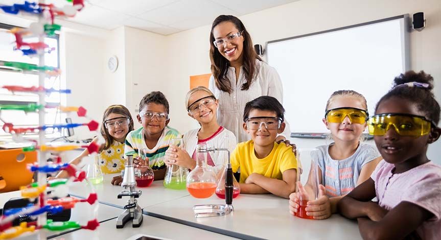 Students wearing safety goggles smiling at the table holding their beakers with multi colored liquid inside them.